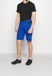 Siniset, kevyestä materiaalista valmistetut shortsit, joissa on tasainen etuosa, sivutaskut ja mustat accentit. Yhdistettynä istuvaan mustaan T-paitaan ja lenkkareihin.