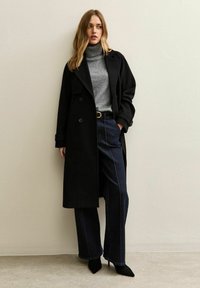 Abrigo negro de doble botonadura, cuello alto gris, pantalones de mezclilla oscuros de talle alto y pierna ancha con costuras visibles, y zapatos de tacón negros puntiagudos.