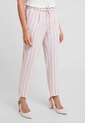 Femme portant un pantalon à rayures verticales roses et blanches avec des chaussures à talons hauts beige à bride arrière sur un fond uni.