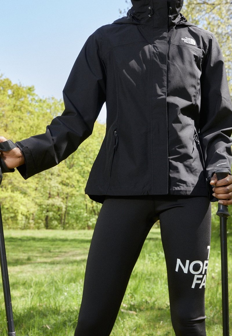 Personne portant une veste noire North Face et des leggings, tenant des bâtons de randonnée, debout en extérieur sur de l'herbe avec des arbres en arrière-plan.