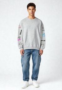 Szürke pulóver hímzett mintákkal, színes szöveggel, laza kék farmer cargo nadrággal és fehér sportcipővel párosítva.