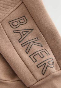 Jasnobrązowa tkanina z teksturowanym dzianiem i wytłoczonym czarnym logo "BAKER". Projekt zawiera miękką, gładką fakturę.