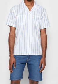 Camisa de manga corta con botones en blanco con rayas verticales azul claro, con dos bolsillos en el pecho y un corte holgado, combinada con pantalones cortos de mezclilla azul.