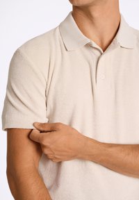 Beigepiké med texturerad tyg, korta ärmar, tre-knappars knäppning och en mjuk krage, visad på en person som justerar ärmen.