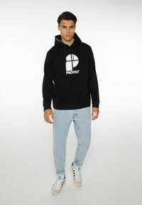 Felpa nera con cappuccio caratterizzata da un logo circolare bianco e la scritta "PROTEST", abbinata a jeans azzurri chiari e sneakers bianche con dettagli neri.