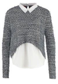 Pull tricoté texturé gris et blanc superposé sur une chemise blanche à col avec manches longues et ourlet asymétrique.
