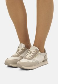 Baskets de sport à lacets avec une palette de couleurs beige et blanche, dotées de tissu texturé, d'accents en daim et de détails en métal doré.