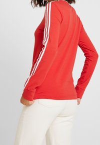 Långärmad röd skjorta i bomull med vita ränder längs ärmarna. Har en figurnära design med rundad hals och ribbade ärmslut.