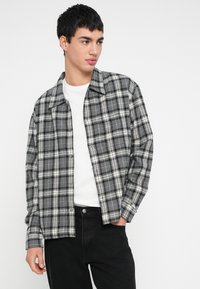 Harmaa ruudullinen flanellipaita, jossa valkoiset ja mustat ruudut, napillinen etuosio ja rento istuvuus. Päällä valkoinen t-paita ja mustat farkut.