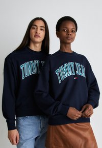 Sudaderas de color azul marino con "TOMMY JEAN" impreso en blanco y verde azulado. Con puños y dobladillo acanalados, usadas con jeans y falda marrón. Tejido de mezcla de algodón suave.