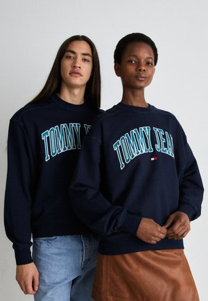 Sudaderas de color azul marino con "TOMMY JEAN" impreso en blanco y verde azulado. Con puños y dobladillo acanalados, usadas con jeans y falda marrón. Tejido de mezcla de algodón suave.