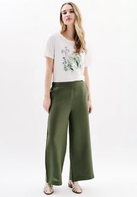 Camiseta blanca con un gráfico colorido de flores y animales, combinada con pantalones de pierna ancha en color verde olivo y sandalias de tiras en un tono beige claro.