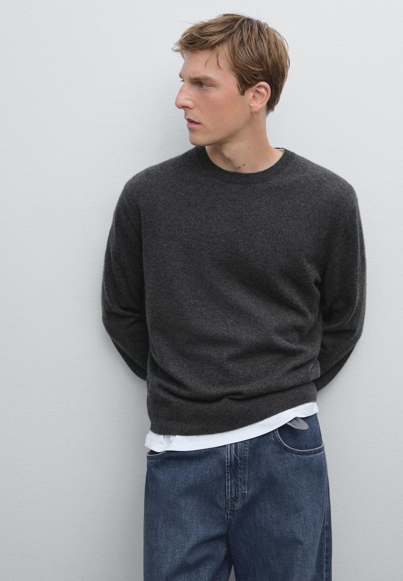 Czarny sweter z okrągłym dekoltem wykonany z miękkiego materiału, noszony na białej koszuli, w połączeniu z niebieskimi dżinsami. Prosty design z długimi rękawami.