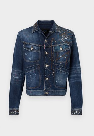 Νέος άνδρας που φοράει τζιν μπουφάν με καρφιά, λευκό μπλουζάκι, καμουφλάζ cargo σορτς με μπαλώματα, λευκές κάλτσες και μαύρα αθλητικά, στέκεται μπροστά σε απλό φόντο.