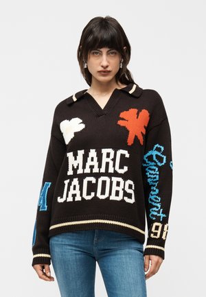 Sötéthajú nő fekete kötött pulóverben, amelyen a "MARC JACOBS" felirat, virágminták és kék írott szöveg található a jobb ujjon, kék farmernadrággal párosítva.