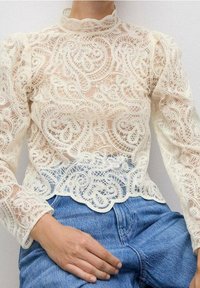Blouse en dentelle crème avec un motif paisley complexe, col haut et manches bouffantes. Associée à un jean taille haute.