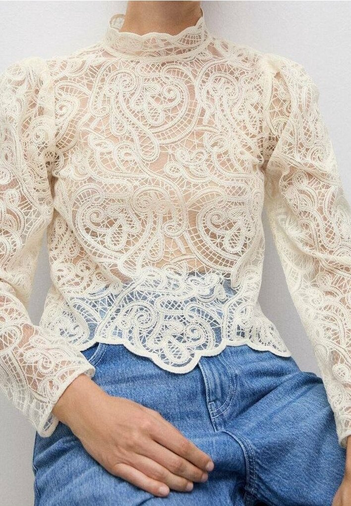 Blouse en dentelle crème avec un motif paisley complexe, col haut et manches bouffantes. Associée à un jean taille haute.