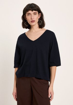 Woman with short dark hair wears a loose black V-neck top, brown pants, and large geometric square earrings, standing against a plain light background.