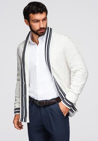 Cardigan bianco lavorato a maglia con un motivo a treccia e colletto a scialle a righe blu navy, indossato sopra una camicia bianca e pantaloni scuri.