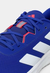 Blå sportsko med hvite striper, blå lisser, rød og grå aksent på tungen, og teksturert mesh-overdel.