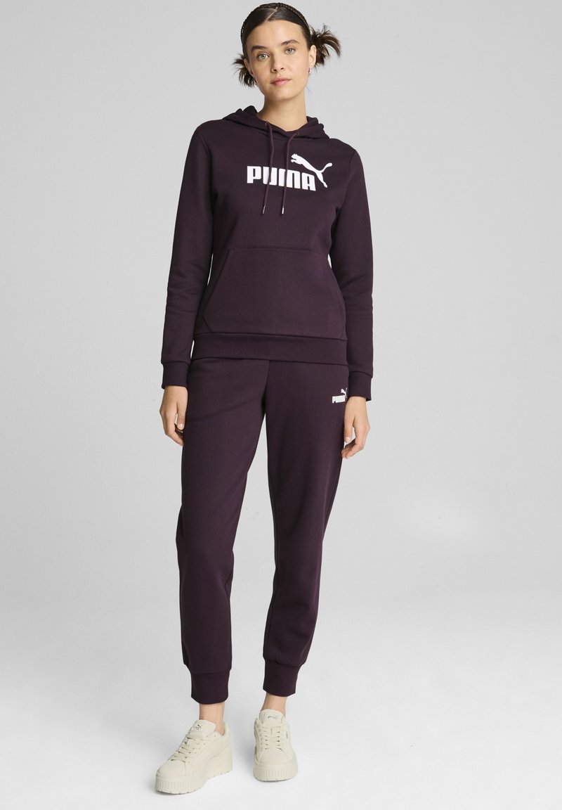 Tuta di colore viola scuro con felpa con cappuccio che presenta un logo bianco, abbinata a pantaloni jogger coordinati. Tessuto morbido, polsini a coste. Sneakers bianche.