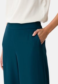 Teal, tailored trousers with a smooth fabric texture, featuring side pockets and a flat waistband, paired with a white blouse.