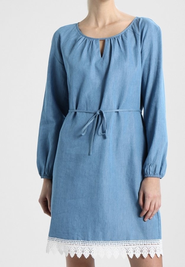 Femme portant une robe bleu clair à manches longues avec une ceinture à la taille et une bordure en dentelle blanche à l'ourlet, debout devant un fond uni.
