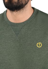 Olive green sweatshirt with a ribbed crew neckline, textured fabric, and a small yellow circular logo embroidered on the chest.