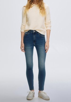 Woman wearing a cream long-sleeve polo sweater, blue skinny jeans, and white sneakers, standing against a plain light background.