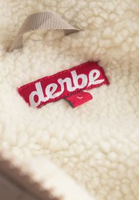 Tissu doux de couleur crème avec une texture duveteuse ; présente une étiquette rouge proéminente portant le mot "derbe" en lettres blanches. Petite boucle grise en accent.