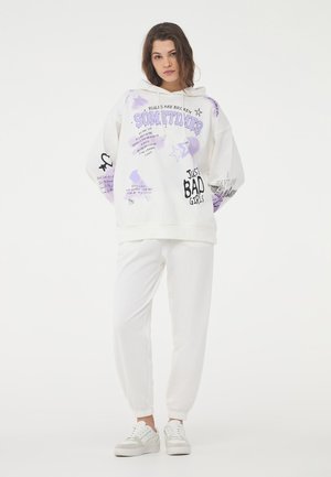 Hoodie bianco oversize con stampe grafiche viola, coulisse e dettagli testuali. Abbinato a pantaloni jogger bianchi e sneakers bianche con accenti grigi.