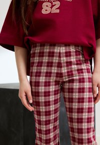 Weinrote Oversize-Hemd mit weißem Aufdruck, kombiniert mit hoch taillierter karierten Hose in Weinrot und Beige. Glatte Textur mit kariertem Muster.