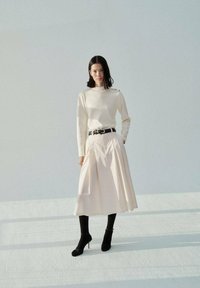 Cream sweater with a high neck, paired with a pleated light pink skirt. Black belt and ankle boots complete the look. Smooth textures, minimalist design.