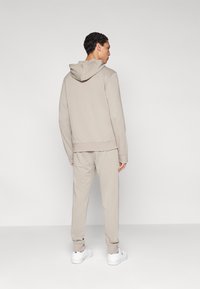 Μπεζ φούτερ με κουκούλα και παντελόνι στο ίδιο χρώμα από απαλό ύφασμα. Διαθέτει ελαστικά τελειώματα στα μανίκια και στη μέση, με μινιμαλιστικό σχεδιασμό και ουδέτερο τόνο.