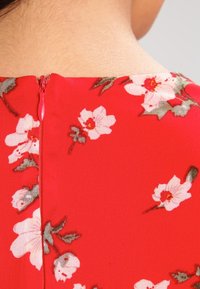 Tela roja con un patrón floral que presenta acentos en blanco y verde, mostrando un cierre de cremallera en la parte posterior cerca del escote.