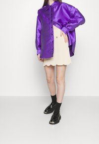 Chemise surdimensionnée en satin violet, jupe beige à bord festonné, et chaussures noir verni à semelles épaisses. La tenue présente des boutons et des textures brillantes.
