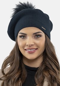 Beret negru din tricot cu o panglică pe vârf, decorat cu un accent metalic mic. Purtați de o persoană cu păr lung și ondulat.