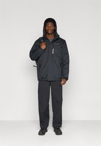 Jachetă neagră izolată cu glugă, buzunare cu fermoar și manșete ajustabile, purtată cu pantaloni cargo negri asortați și pantofi negri.