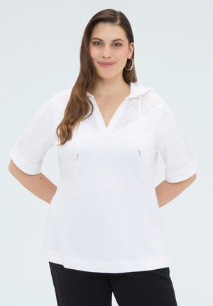 Frau mit langen braunen Haaren, die Creolen trägt, einen weißen kurzärmeligen Hoodie mit Spitzen-Details und schwarzen Hosen, stehend mit den Händen hinter dem Rücken.