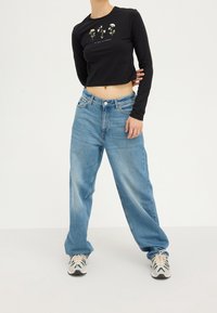 Camisa negra de manga larga corta con gráficos florales, combinada con jeans de corte holgado en un tono azul claro y zapatillas deportivas de tonos neutros.