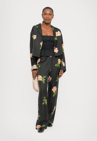 Blazer cu model floral negru și pantaloni evazați cu trandafiri piersici pe un fundal întunecat, asortați cu un top texturat negru și o geantă bej.