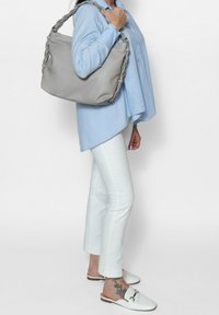 Borsa a mano in pelle grigia con manico intrecciato, texture morbida, design spazioso. Indossata sopra una camicia azzurro chiaro e pantaloni bianchi.