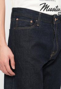 Κοντινό πλάνο σκούρου μπλε τζιν με κίτρινη ραφή, φορεμένα με λευκή μπλούζα με το λογότυπο Mustang, δείχνοντας το αριστερό χέρι και τις μπροστινές τσέπες.