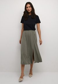 Zwart T-shirt met korte mouwen gecombineerd met een vloeiende, olijfgroene maxi-jurk met een split aan de voorkant, aangevuld met beige instapschoenen.
