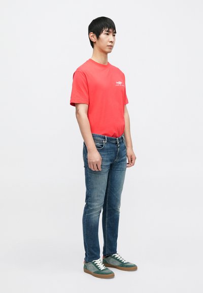 T-shirt en coton rouge avec un petit motif graphique, assorti à un jean en denim bleu et des baskets en daim vert avec des lacets blancs et des semelles marron.