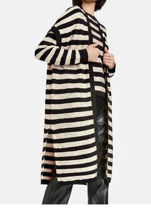 Femme portant un long cardigan rayé horizontal noir et beige par-dessus un pull assorti et un pantalon en cuir noir, tenant les bords du cardigan.
