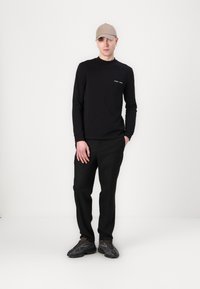 Maglietta nera a maniche lunghe con piccolo testo bianco, abbinata a pantaloni neri e sneakers grigie. La persona indossa un cappellino beige chiaro.