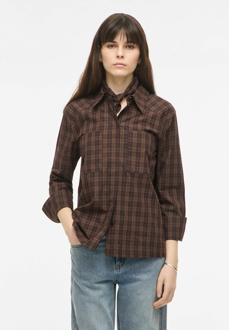 Jeune femme aux longs cheveux foncés portant une chemise à carreaux marron et un jean bleu clair, debout avec une main dans sa poche.