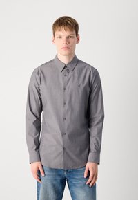 Camisa de manga larga a rayas grises con collar abotonado, que presenta un logotipo sutil en el pecho y botones marrones. Combinada con vaqueros de mezclilla azules.