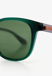 Zielone plastikowe okulary przeciwsłoneczne z ciemnymi soczewkami, z pomarańczowym brandingiem "SUPERDRY" na zausznikach i gładkim matowym wykończeniem.